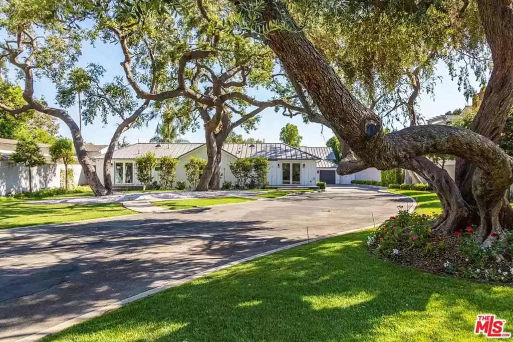 Past grey gates and knobbly oak trees, a motor court reveals a white farmhouse-style compound with a grey roof.